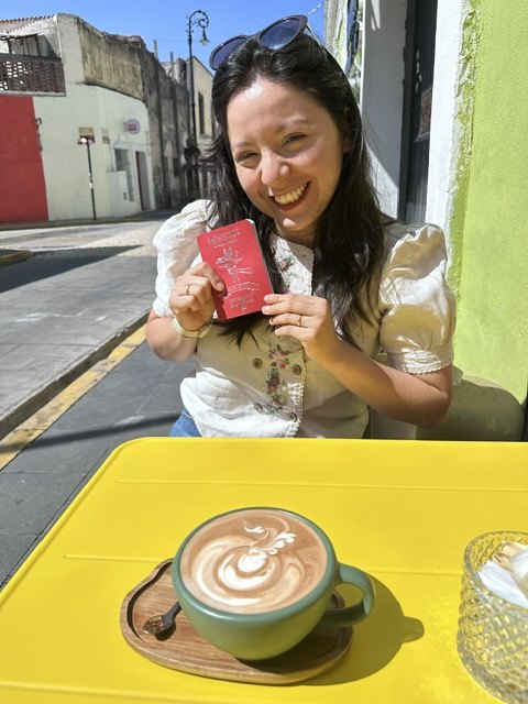 Diana holding the coffee passport at a yellow table on a colourful Atlixco street