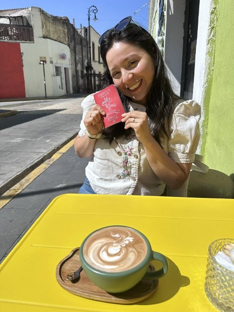 Diana holding the red Puebla coffee passport at a yellow table outside Cuac Café in Atlixco