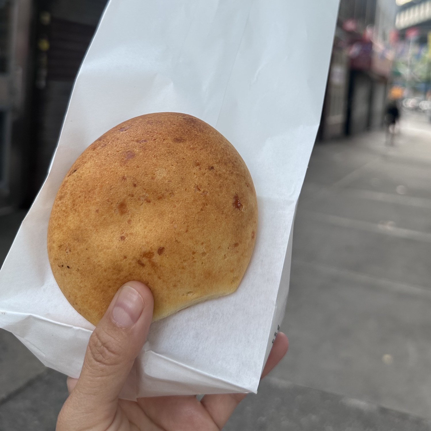 A golden pan de bono held on white bakery paper outside Rico Pan Bakery in Woodside, Queens